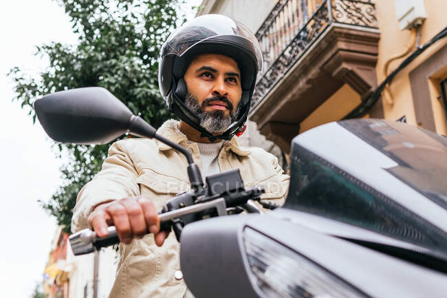 Von unten der brutale ethnische männliche Biker mit Helm, der zeitgenössisches Motorrad fährt, während er sich auf die Stadt freut — Stockfoto
