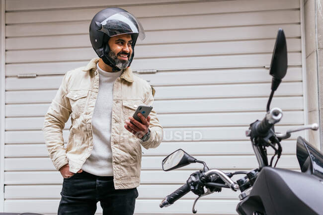 Content mature ethnic male motorcyclist with cellphone and hand in pocket looking away against bike in town — Stock Photo