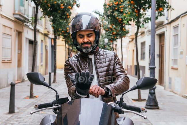 Mature bearded Hispanic male biker in protective helmet putting on glove on contemporary motorcycle on street — Stock Photo