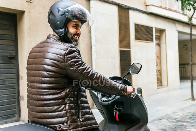 Side view of brutal ethnic male biker in helmet driving contemporary motorbike while looking way in town — Stock Photo