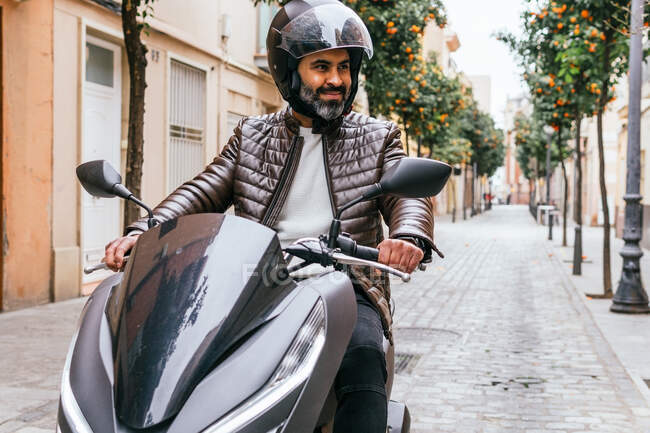 Жорстокий етнічний чоловік - байкер у шоломі їздить на сучасному мотоциклі, дивлячись далеко в місті — стокове фото