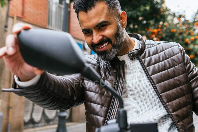 Crop glad middle aged ethnic male motorcyclist with beard looking in side mirror on bike in city — Stock Photo
