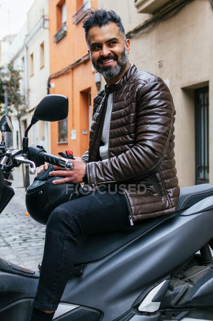 Vista lateral del contenido masculino hispano motociclista masculino con casco mirando hacia otro lado en moto en la ciudad - foto de stock