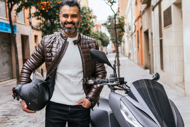 Content masculine Hispanic male motorcyclist with helmet looking away standing near motorbike in town — Stock Photo