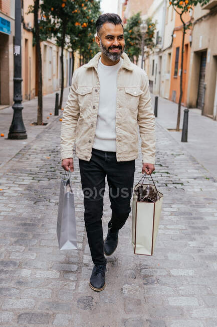 Heureux homme ethnique d'âge moyen avec des sacs à provisions se promener sur le trottoir urbain et regarder loin — Photo de stock