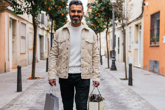 Feliz hombre étnico de mediana edad con bolsas de compras paseando por la pasarela urbana y mirando a la cámara - foto de stock