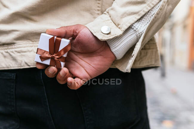 Back view of crop anonymous male with hand behind back holding small present box with ribbon in town — Stock Photo