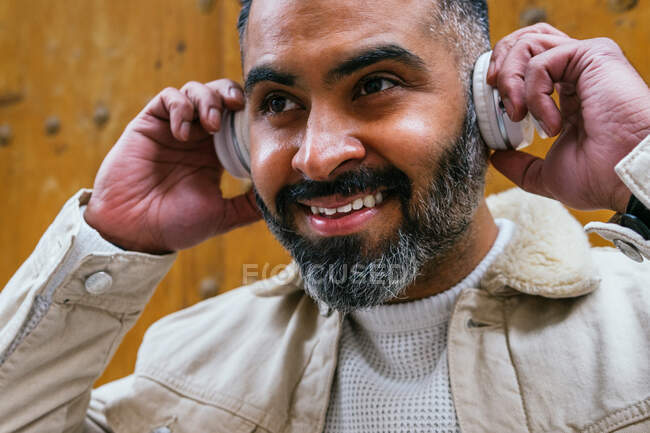 Обрізати доброзичливий бородатий етнічний чоловік слухає пісню з бездротового гарнітура, дивлячись вдень — стокове фото