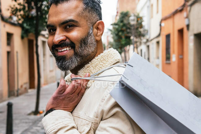 Side view of crop smiling masculine Hispanic man with gift bag looking away in city — Stock Photo