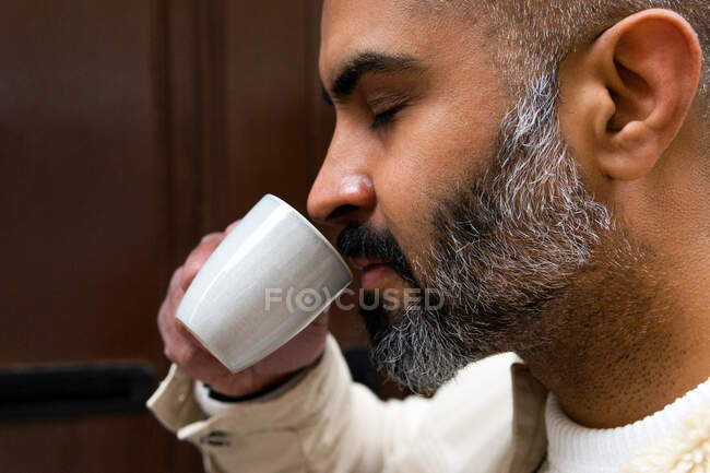 Vista lateral de la cosecha concentrada de mediana edad étnica masculina con ojos cerrados disfrutando del aroma de la bebida caliente en la taza - foto de stock