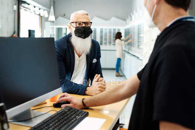 Elderly male in mask standing at desk and talking to optician in optical store — Stock Photo