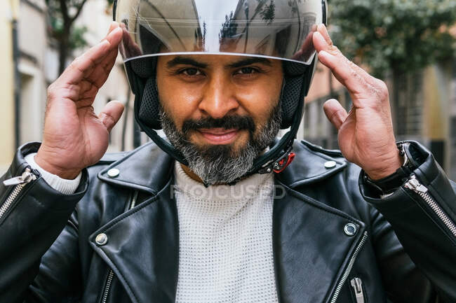 Cheerful middle aged ethnic male motorcyclist putting on protective helmet while looking at camera against ribbed wall — Stock Photo