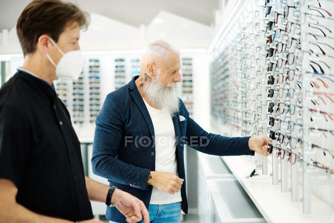 Side view of optician in mask and senior male choosing eyeglasses in optical store — Stock Photo