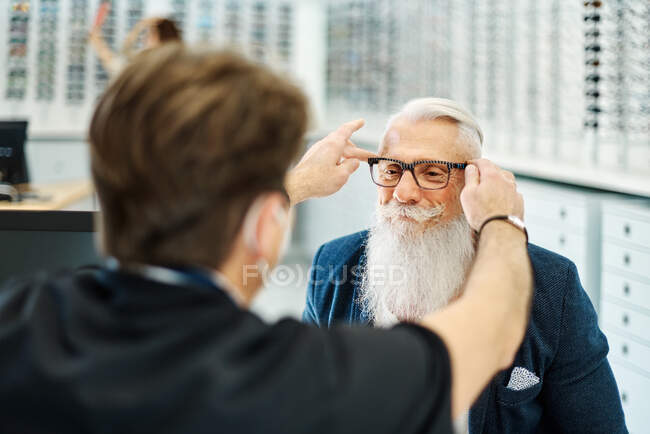 Обратный вид профессионального оптика в маске, помогающего пожилым мужчинам надевать очки во время работы в оптическом магазине — стоковое фото