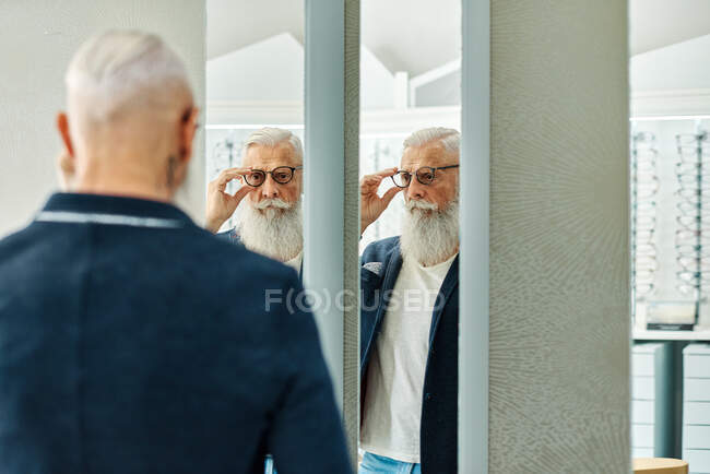 Вид сзади на стильного мужчину, стоящего перед зеркалом в оптическом магазине и примеряющего модные очки — стоковое фото