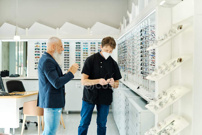 Side view of professional optician in mask helping senior male choosing glasses while working in optical store — Stock Photo