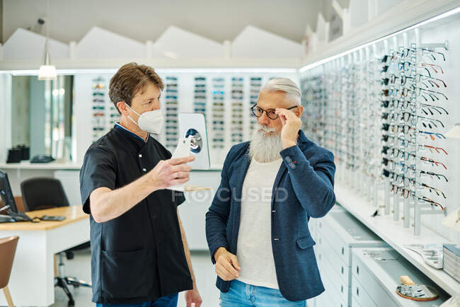Side view of professional optician in mask helping senior male choosing glasses while working in optical store — Stock Photo