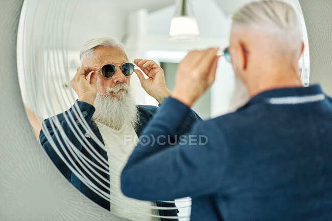 Vista trasera de un hombre mayor con estilo de pie frente al espejo en la tienda óptica y probándose gafas de sol de moda - foto de stock