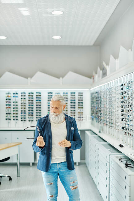Serious elderly bearded male in trendy outfit standing with modern stylish glasses in optical shop — Stock Photo