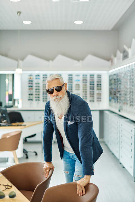 Самоуверенный пожилой мужчина в модных солнцезащитных очках и одежде, стоящий в оптическом магазине и смотрящий в камеру — стоковое фото