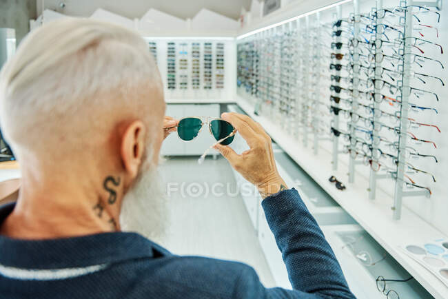 Back view elderly bearded male in trendy outfit standing with modern stylish sunglasses in optical shop — Stock Photo