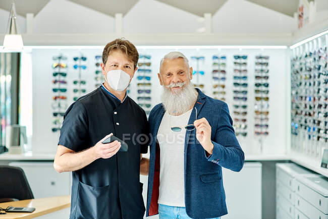 Улыбающийся пожилой мужчина-клиент и оптик, стоящий с очками в оптическом магазине и смотрящий в камеру — стоковое фото