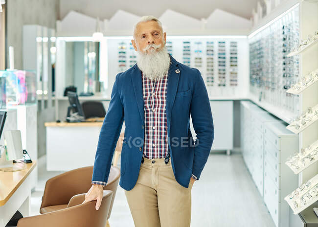 Determined elderly male hipster in trendy outfit standing in modern optical store and looking away — Stock Photo