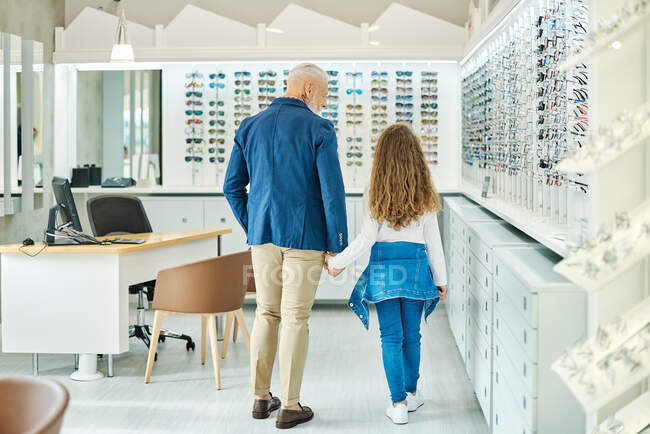 Вид сзади на стильного пожилого мужчину, держащего за руку внучку-подростка, стоя в оптическом магазине — стоковое фото