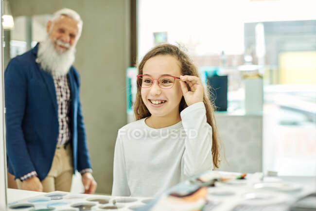 Feliz adolescente probándose las gafas mientras está de pie en la tienda óptica con el abuelo y mirando en el espejo - foto de stock