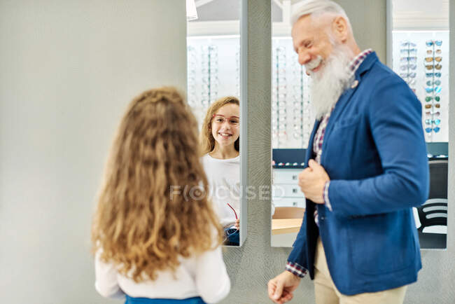 Vista trasera de la adolescente probándose las gafas mientras está de pie en la tienda óptica con el abuelo y mirando en el espejo - foto de stock