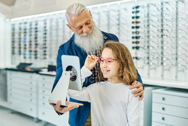 Fröhlicher älterer Mann hält Spiegel, während Teenager-Mädchen Brille in modernem Optikgeschäft anprobiert — Stockfoto