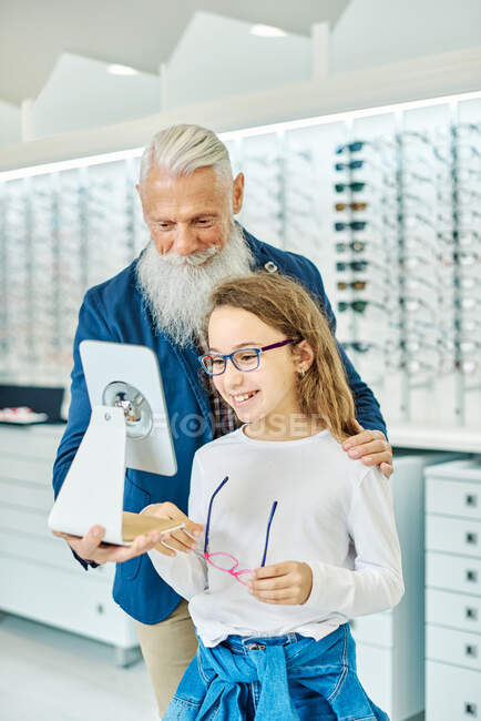Allegro uomo anziano che tiene lo specchio mentre l'adolescente si prova gli occhiali nel moderno negozio ottico — Foto stock