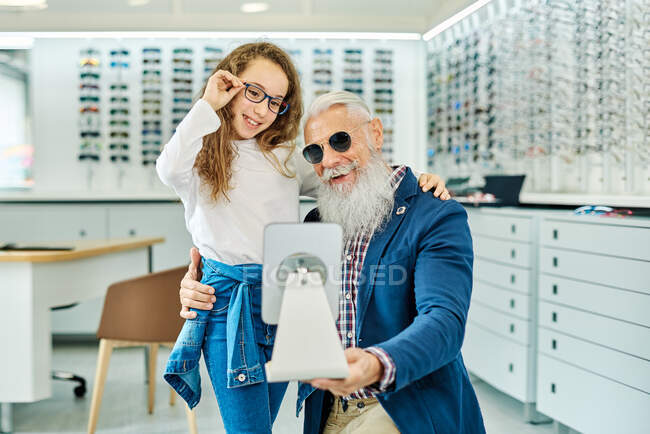Веселый пожилой мужчина держит зеркало, пока девочка-подросток примеряет очки в современном оптическом магазине — стоковое фото