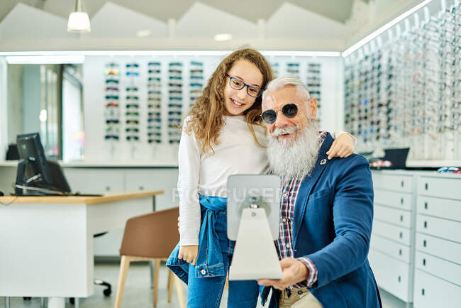 Веселый пожилой мужчина держит зеркало, пока девочка-подросток примеряет очки в современном оптическом магазине — стоковое фото