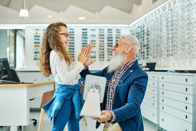 Side view of cheerful grandfather and granddaughter in glasses giving high five to each other in optical store — Stock Photo