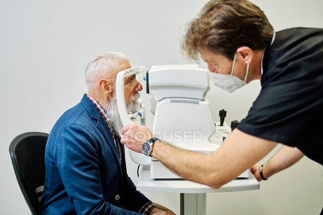 Vista lateral do paciente sênior do sexo masculino sentado na cadeira na frente do tonômetro moderno para medir a pressão ocular na clínica com oftalmologista — Fotografia de Stock