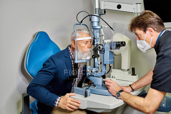 Vista lateral do oftalmologista profissional usando lâmpada de fenda e verificação da visão do paciente do sexo masculino sênior — Fotografia de Stock