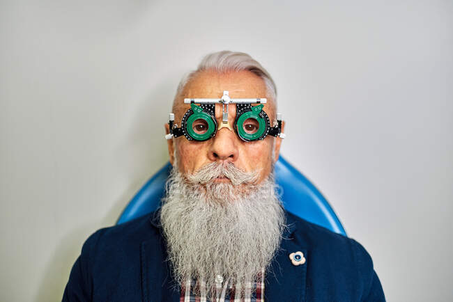 Bearded elderly male in eye test glasses sitting in modern clinic before vision examination and looking at camera — Stock Photo