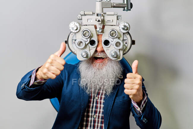 Feliz macho mayor con foropter durante el examen ocular mirando a la cámara y el pulgar hacia arriba - foto de stock