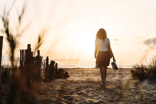 Обратный вид на неузнаваемую женщину в летней одежде идущую к морю вдоль песчаного пляжа на фоне закатного неба — стоковое фото