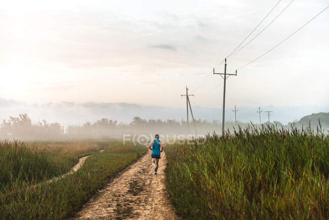 Вид ззаду невпізнаваний чоловічий бігун біжить піщаним шляхом у сільській місцевості під час тренувань в туманний ранок влітку — стокове фото