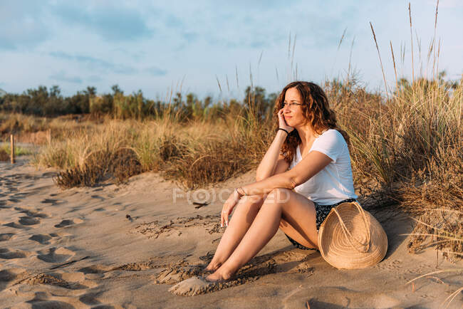 Vue latérale d'une femme tranquille assise sur un rivage sablonneux et penchée sur la main tout en admirant le coucher du soleil en été — Photo de stock