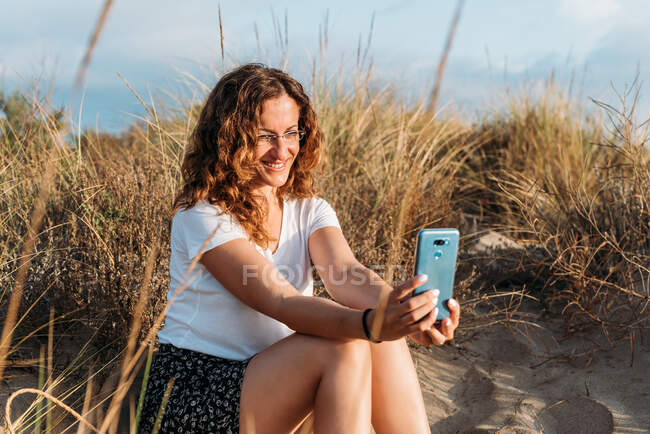 Contenida hembra en traje de verano sentada en la orilla arenosa y tomando selfie con smartphone mientras disfruta de la puesta de sol - foto de stock