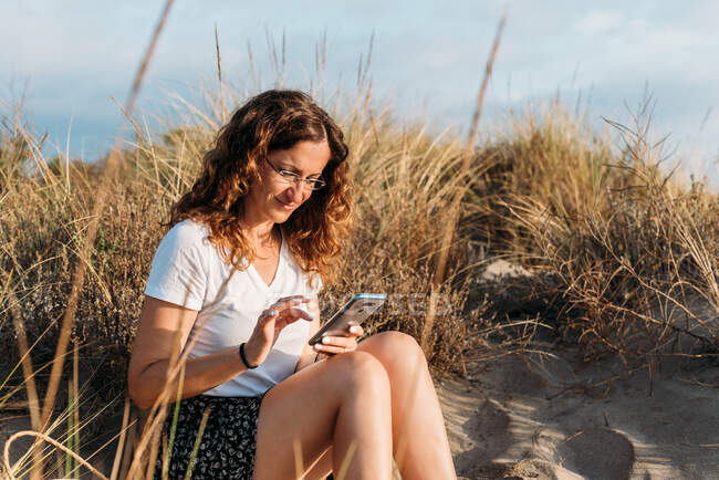 Zufriedene Frau im Sommer-Outfit sitzt am Sandstrand und sendet Nachrichten auf dem Smartphone, während sie den Sonnenuntergang genießt — Stockfoto