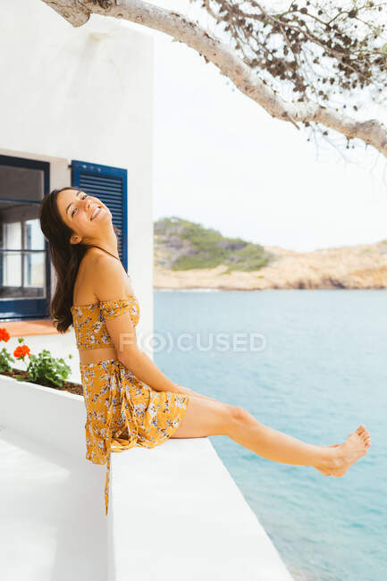 Вид збоку весела молода жінка в літній сукні, що сидить на терасі заміського будинку, розташованого на мальовничому березі озера і насолоджується літнім відпочинком — стокове фото
