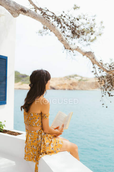Side view of romantic young female in summer outfit sitting on terrace near lake water and reading interesting novel in book while spending summertime in countryside — Stock Photo