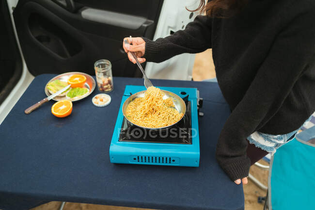 High angle side view of unrecognizable female traveler preparing spaghetti in frying pan on portable gas stove near camping van — Stock Photo