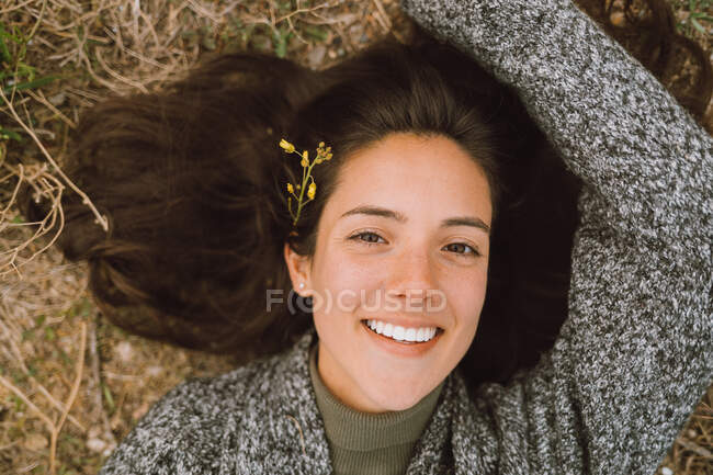 Top view of cheerful young female in warm knitted wear with blooming flower in hair looking at camera while enjoying spring day during travel in nature — Stock Photo