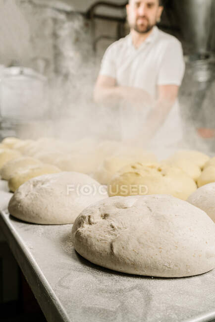 Crop ethnic male baker in uniform at table with raw bread dough and flour dust in bakery — Stock Photo