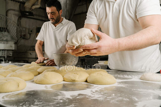 Cropped mature bearded ethnic bakers forming bread from dough at table with flour and bowl in bakery — Stock Photo